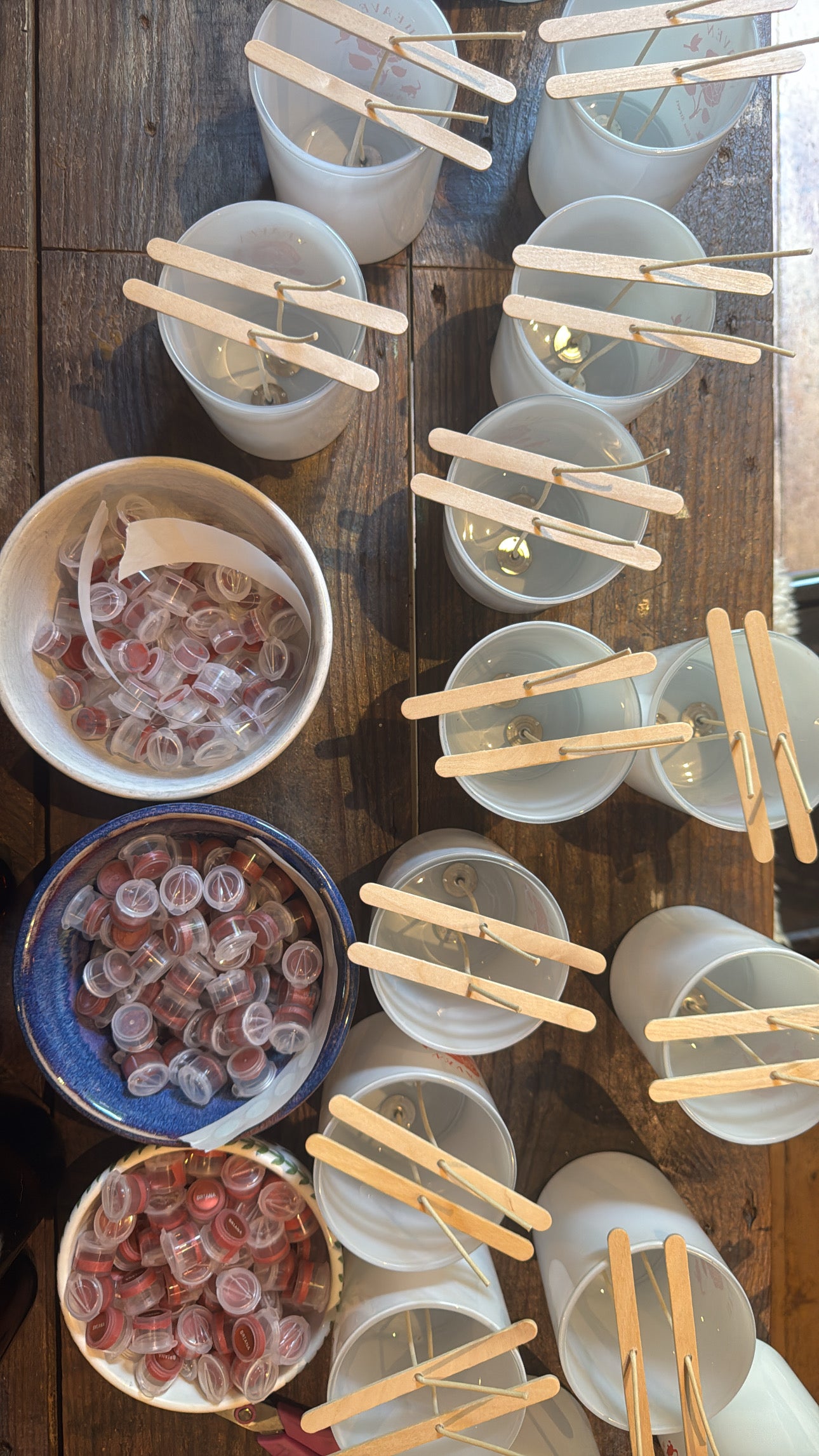 Clear plastic cups with wooden sticks on a wooden surface, with bowls of pink and white objects nearby.