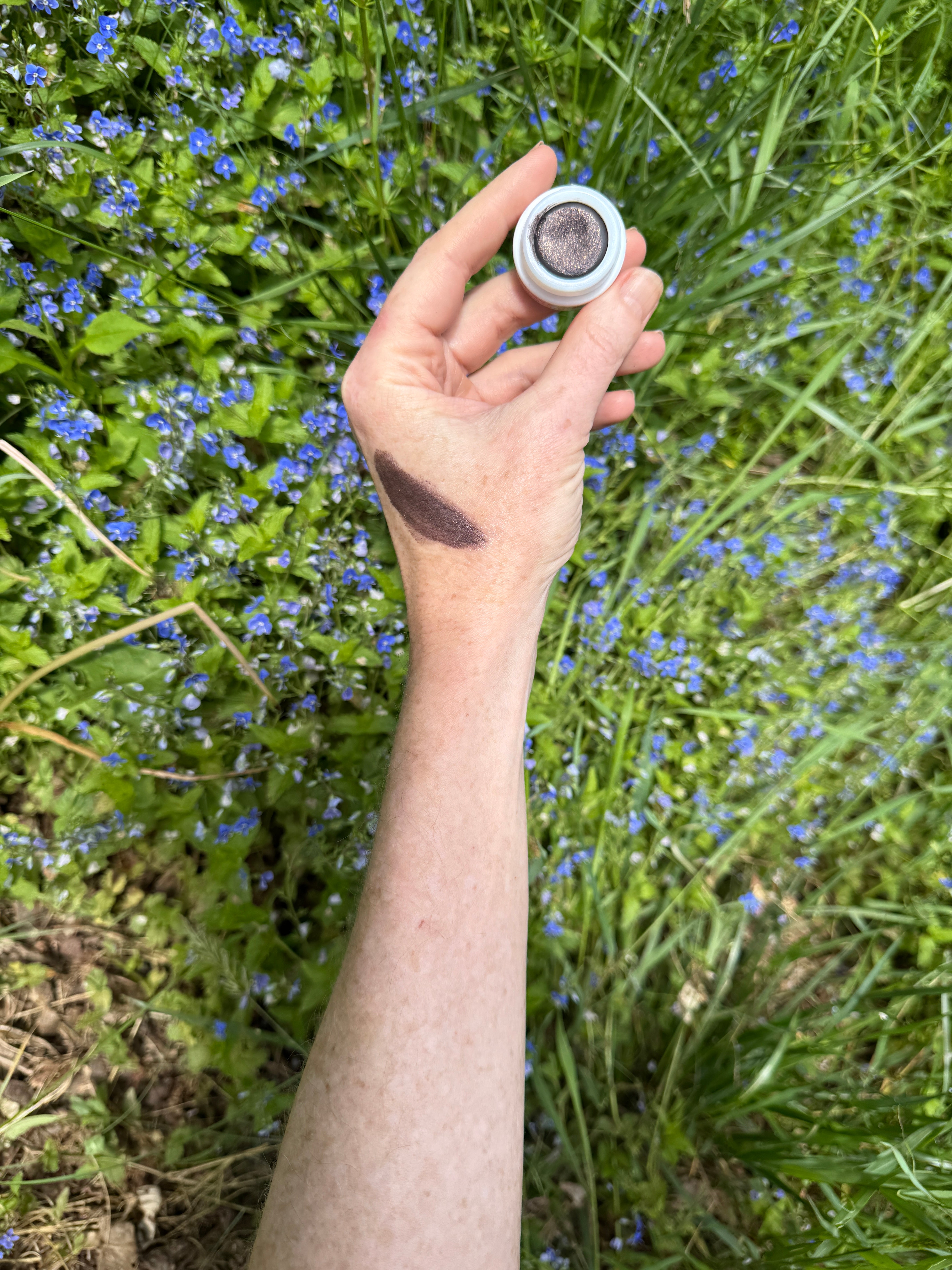 Hand holding a small white container with a dark substance against a natural background of green grass and blue flowers.