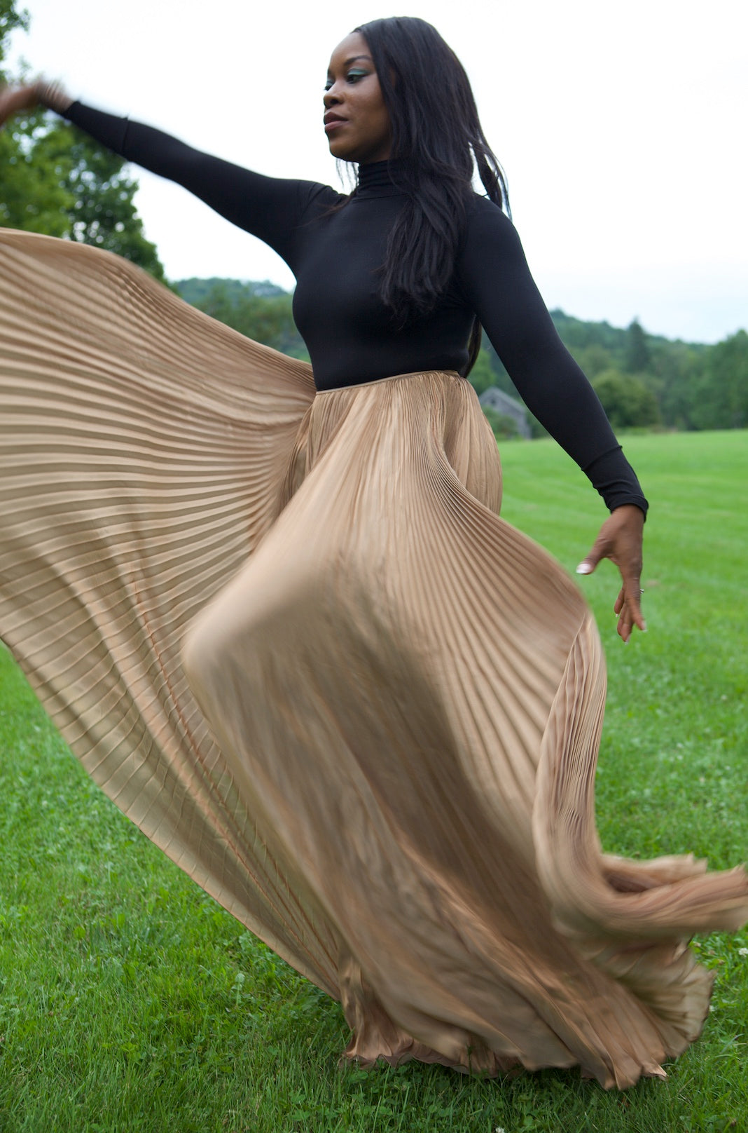 Woman in a black top and long beige skirt standing on grass with trees in the background